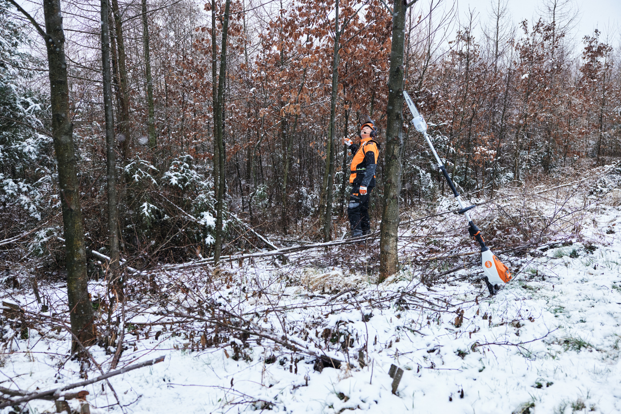 En man i skyddsutrustning arbetar i en snöig skog med en STIHL batteridriven stamkvistare HTA 150 och lutar sig mot en trädstam i förgrunden