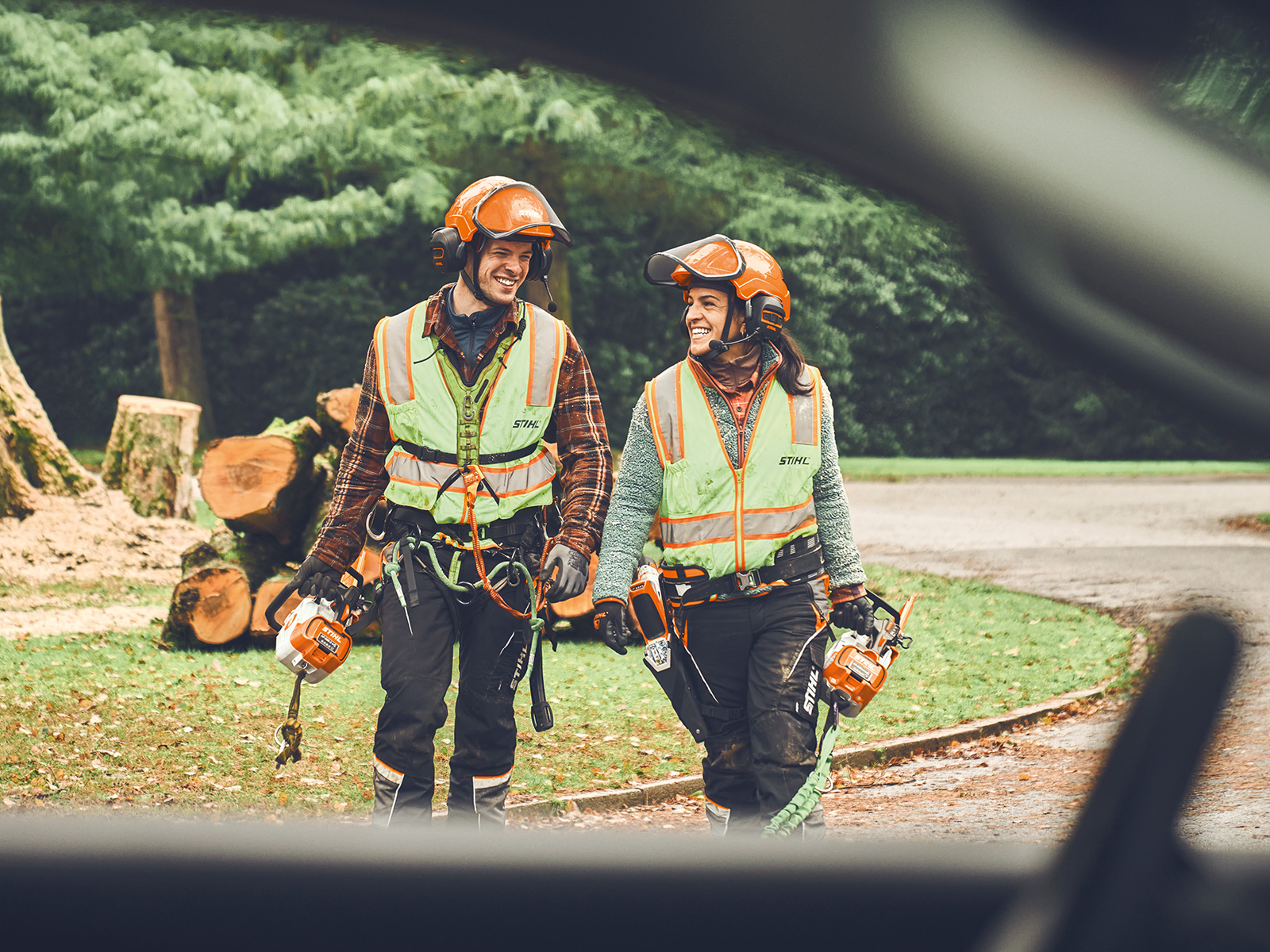 En man och en kvinna med STIHL sladdlösa motorsågar och en STIHL sladdlös beskärare går leende mot en bil  