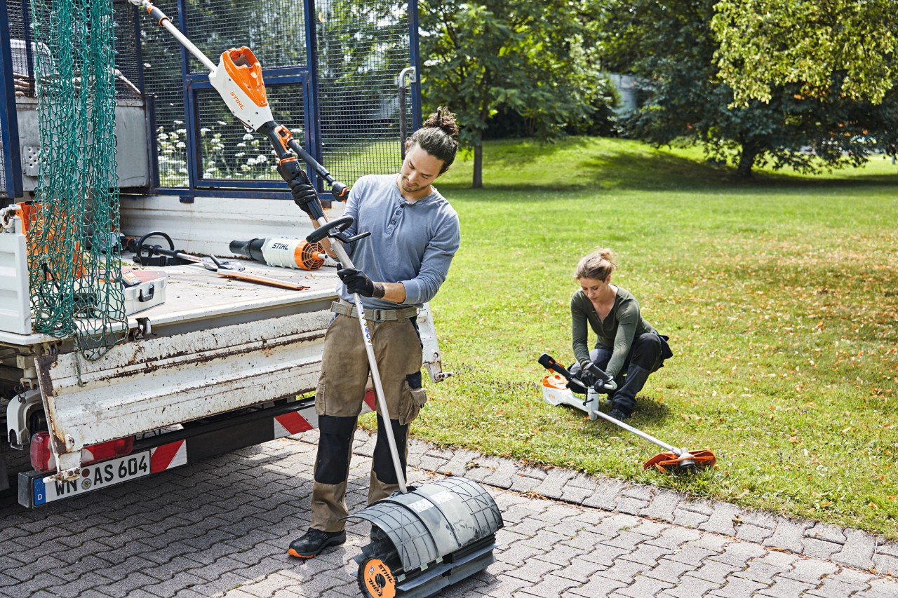 En man och en kvinna hanterar STIHL batteridrivna maskiner intill ett fordon med öppen bagelucka, en äng i bakgrunden
