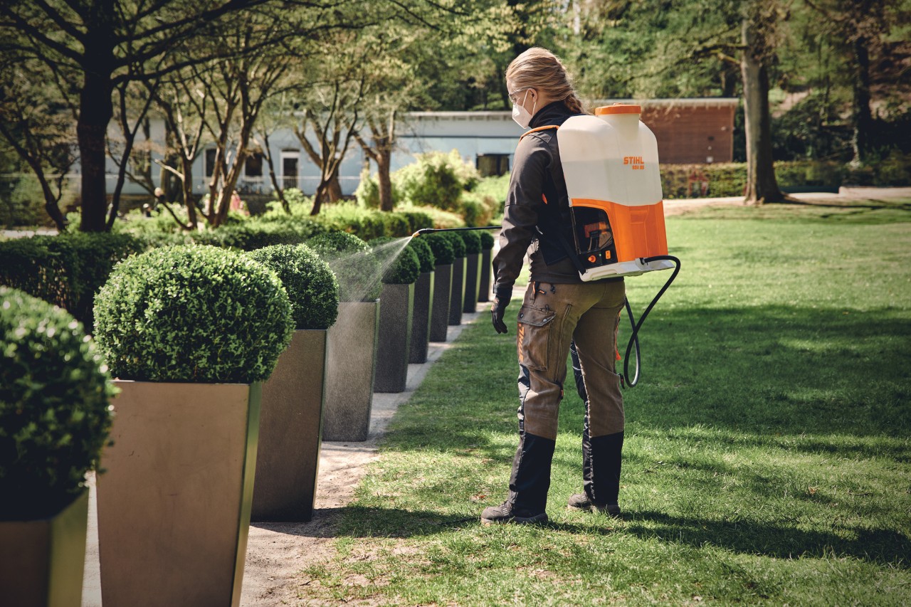 Eine blonde Person mit Mundschutz wässert mit einem STIHL Spritzgerät auf einer Grünfläche rund geschnittene Büsche, die einen Weg säumen